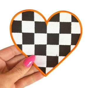 May include: Heart-shaped patch with a black and white checkered pattern. The patch has an orange border and is held against a white background. The patch is approximately 3 inches wide.