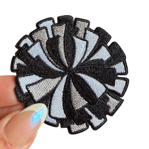May include: A round, embroidered patch with a radial design. The patch features alternating segments of black, light blue, and gray thread, creating a textured, dimensional effect. The patch is held by a hand against a white background.