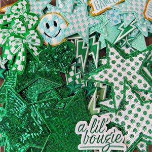 May include: A collection of green and white embroidered patches in various shapes, including stars, bows, hearts, and lightning bolts. Some patches feature sequins, checkered patterns, and the text "a lil bougie".