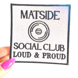 May include: Square white embroidered patch with black text and a circular graphic. The text reads "MATSIDE SOCIAL CLUB LOUD & PROUD". The patch is held by a hand with a pink painted fingernail.