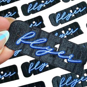 May include: Close-up of several black fabric patches with the word "flyer" embroidered in blue. The patches have a textured surface and a rectangular shape with rounded corners. Some patches are held by a hand.