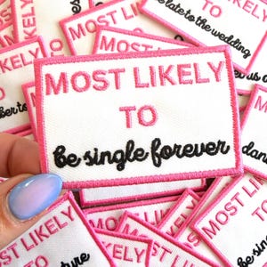 May include: White rectangular patch with pink border and text "MOST LIKELY TO be single forever" in pink and black. Other patches with similar text are in the background.