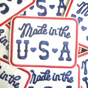 May include: White rectangular patch with a red border and the text "Made in the U.S.A." embroidered in blue. The letters "U", "S", and "A" are separated by hearts. Multiple patches are visible.