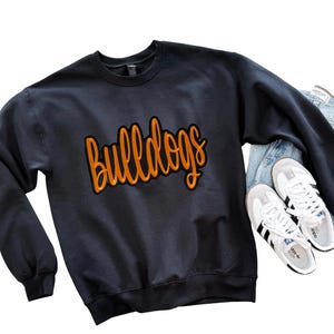 May include: A navy blue sweatshirt with the word "Bulldogs" in orange script. The sweatshirt is paired with blue jeans and white sneakers with black stripes.