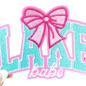 May include: Embroidered patch with the word "LAKE" in turquoise block letters, outlined in pink, and a pink bow. The word "babe" is embroidered in cursive below the word "LAKE".