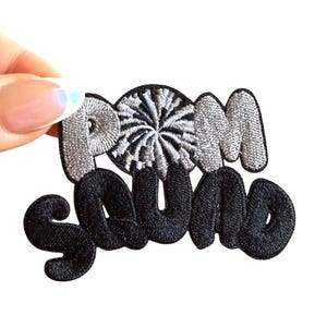 May include: Embroidered patch with the words "POOM SQUAD" in a stylized font. The word "POOM" is in silver thread with a central circular design, and "SQUAD" is in black thread. The patch is held against a white background.