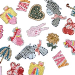 May include: Assorted embroidered patches in various shapes and colors. Designs include a camera with a rainbow, a peace sign, a heart, a flower, a gun with a pink bow, cherries, and the word "YA".