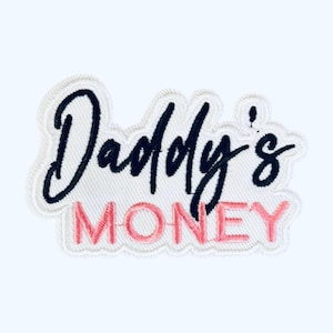 May include: Embroidered patch with the words "Daddy's MONEY" in black and pink thread. The text is on a white background with a decorative border. The patch is likely for clothing or accessories.