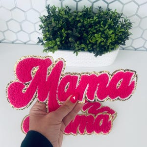 May include: Pink felt "Mama" letters with gold glitter trim. The letters are in a cursive font and are slightly raised.