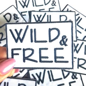 May include: White rectangular patch with the words "WILD & FREE" embroidered in dark blue. The patch has a dark blue border. Several similar patches are visible in the background.