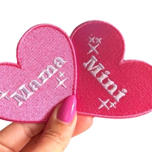 Puede incluir: Dos parches bordados en forma de corazón rosa. Uno dice "Mama" en escritura blanca, y el otro dice "Mini". Ambos parches tienen detalles de estrellas. Los parches son sostenidos por una mano con esmalte de uñas rosa.