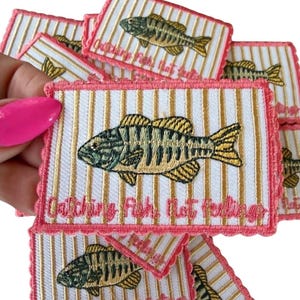 May include: Embroidered patches with a fish design. Each patch features a green and yellow fish on a white background with gold vertical stripes. The patches have a pink scalloped border and the text "Catching Fish, Not Feelings".