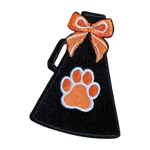 May include: Embroidered black megaphone patch with an orange bow and paw print. The megaphone has a handle and a white outline around the orange paw print. The bow has a white outline.