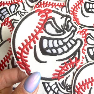 May include: White baseball patch with a cartoon face embroidered in black and red. The baseball has a smiling face with large teeth and red stitching. The patches are round and have a black border.