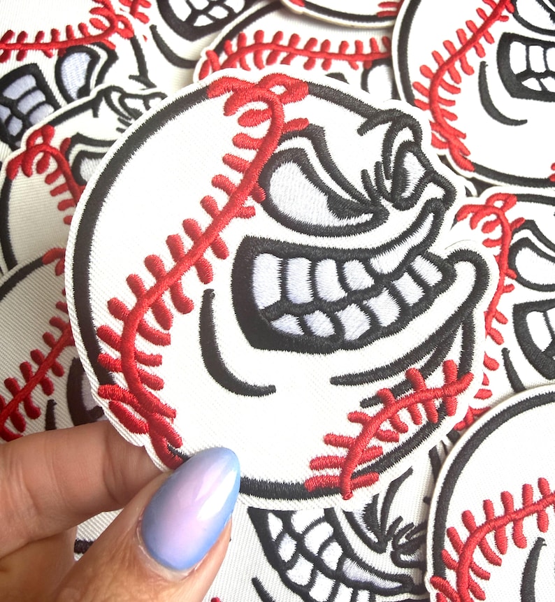 May include: White baseball patch with a cartoon face embroidered in black and red. The baseball has a smiling face with large teeth and red stitching. The patches are round and have a black border.