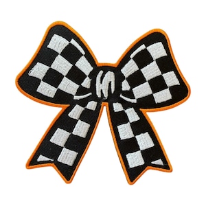 May include: A black and white checkered bow-shaped patch with an orange border. The bow has a stylized design, with the center of the bow featuring a white letter "W".