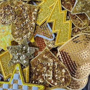 May include: A collection of gold sequin patches in various shapes, including stars, hearts, and lightning bolts. The patches are densely covered in shiny sequins, creating a textured and reflective surface. The patches are likely for embellishing clothing or accessories.