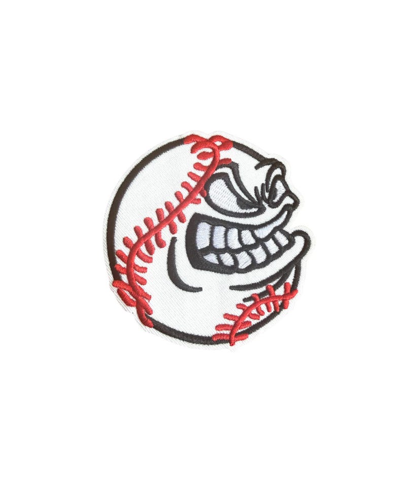 May include: A white baseball patch with red stitching and a black outline. The patch features a cartoon face with a wide, toothy grin, giving it an aggressive appearance. This patch is suitable for sports fans.