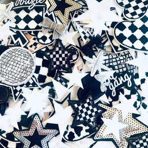 May include: A collection of iron-on patches featuring various designs, including stars, checkered patterns, and the phrase "the gang". The patches are in black, white, and silver.