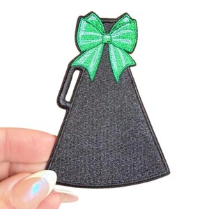 May include: Black megaphone patch with a bright green bow. The patch has a handle and a textured surface. The bow is embroidered with green thread.