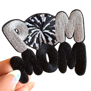 May include: Embroidered patch with the text "POM MOM" in a bubble letter style. The "POM" is silver with black outlining, and the "MOM" is black. A black and white pom-pom design is between the letters.