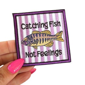 May include: Embroidered patch with a purple border, featuring a fish illustration in yellow, blue, and black. The patch has a striped background and the text "Catching Fish Not Feelings".