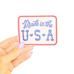 May include: A rectangular patch with a white background, red border, and blue embroidered text that reads "Made in the U.S.A." The letters "U", "S", and "A" each have a small heart symbol. The patch is held by a hand with a light purple manicure.