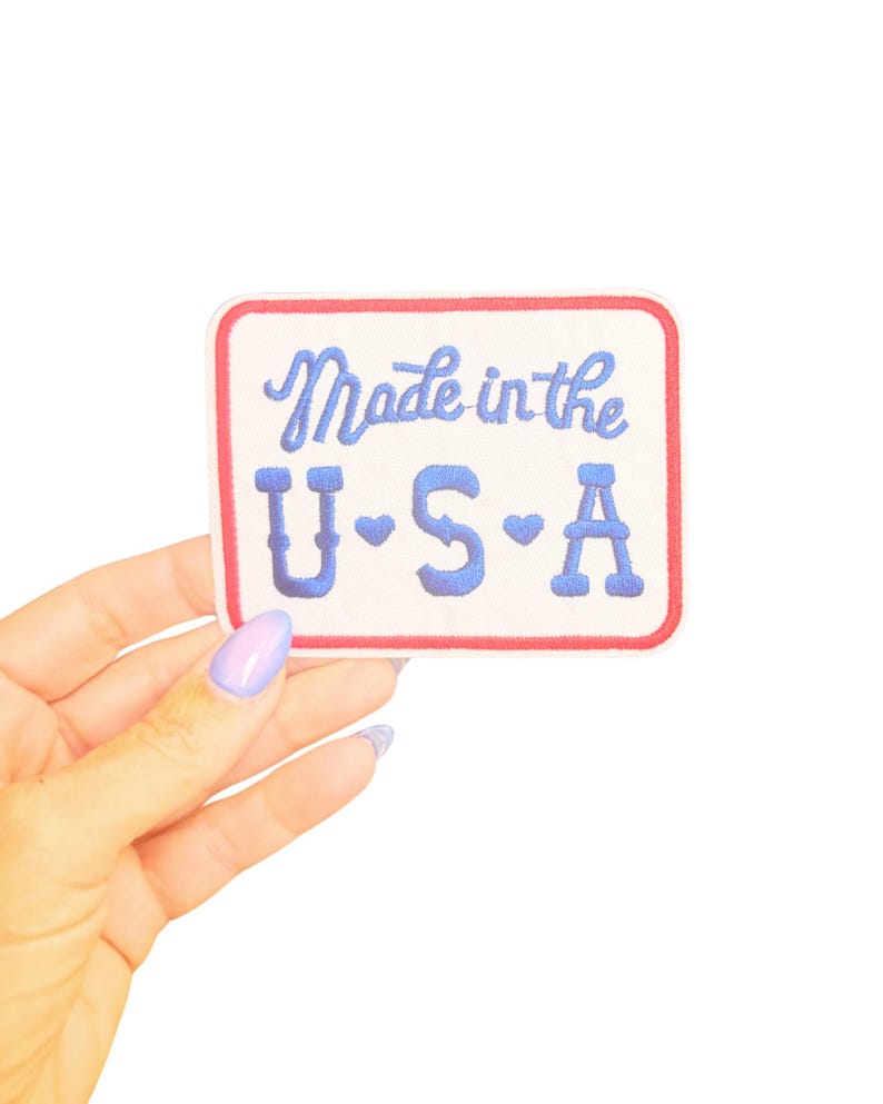 May include: A rectangular patch with a white background, red border, and blue embroidered text that reads "Made in the U.S.A." The letters "U", "S", and "A" each have a small heart symbol. The patch is held by a hand with a light purple manicure.