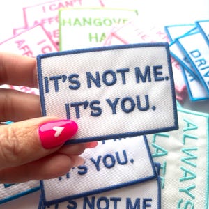 May include: A white fabric patch with blue embroidered text that reads "It's not me. It's you." The patch has a blue border.
