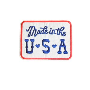 May include: Embroidered patch with a white background, red border, and blue text. The patch reads "Made in the USA" in a cursive font, with "U.S.A" in a block font below. The letters "U" and "S" have blue heart accents.