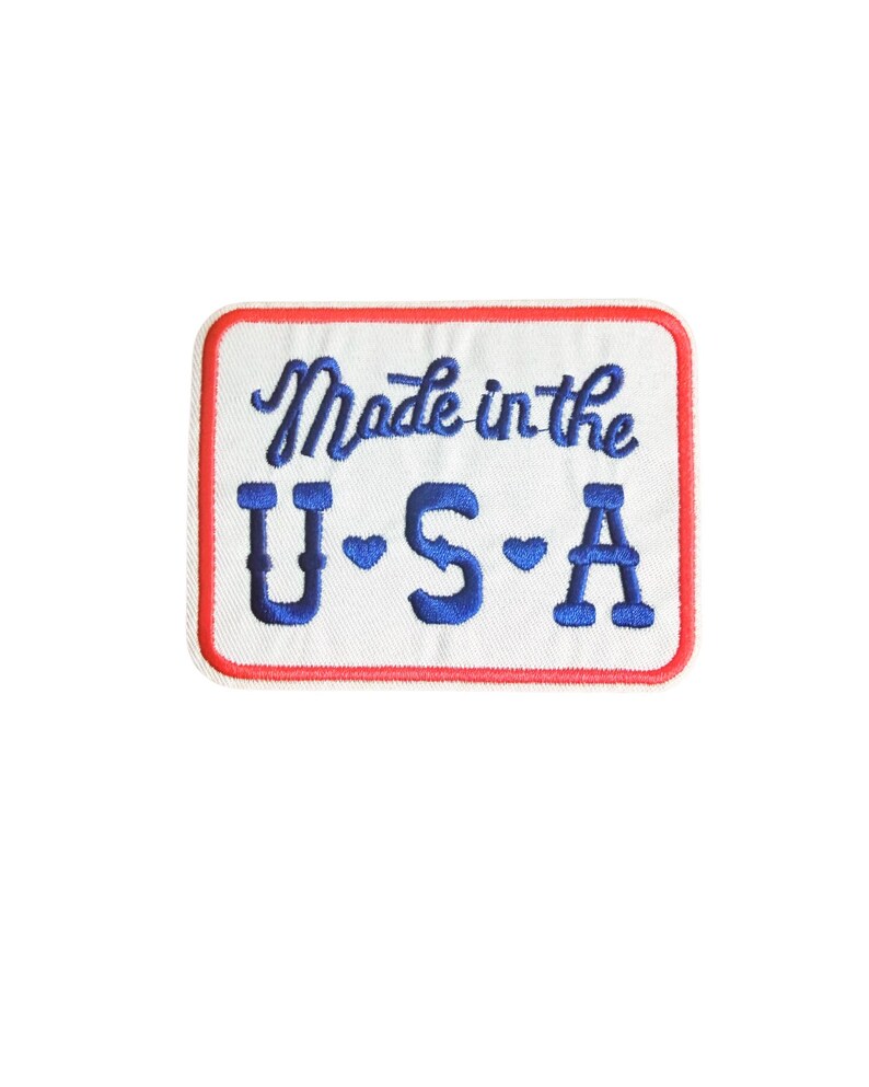 May include: Embroidered patch with a white background, red border, and blue text. The patch reads "Made in the USA" in a cursive font, with "U.S.A" in a block font below. The letters "U" and "S" have blue heart accents.