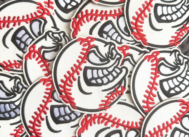 May include: A collection of baseball-themed patches. Each patch features a white baseball with red stitching and a grinning face with black outlines. The patches are scattered across a surface.