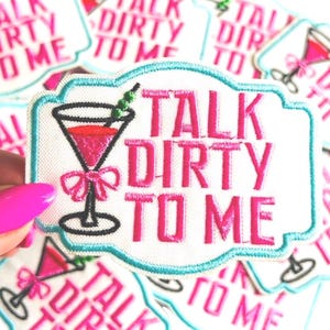 May include: Embroidered patch with the phrase "TALK DIRTY TO ME" in pink lettering. The patch features a pink cocktail glass with a bow and a green olive. The patch has a white background with a light blue border.