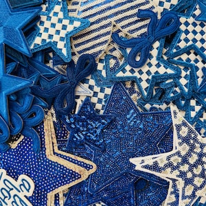 May include: A collection of blue star-shaped patches with various designs. Some stars have sequin details, while others feature checkered or striped patterns. Several patches include bow accents, creating a festive and decorative theme.