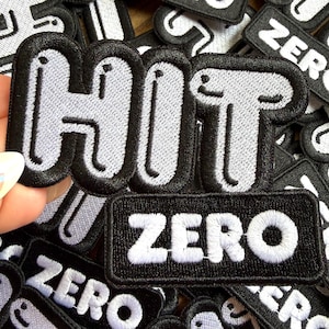 May include: Embroidered patches with the words "HIT" and "ZERO". The "HIT" patch is in a bubble letter style with a black outline and light gray fill. The "ZERO" patch is rectangular with white text on a black background. Patches are black and white.