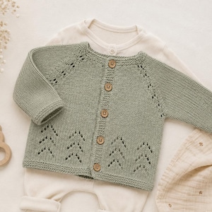 May include: A sage green knitted baby cardigan with wooden buttons and decorative tree-shaped cutouts. The cardigan is laid over a cream-colored onesie and a muslin blanket. A wooden bear-shaped teether is also visible.