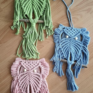 Op de afbeelding: Drie macramé uil wandkleden in groen, blauw en roze. Elke uil heeft een gedetailleerd geweven ontwerp, een houten zitstok en witte kralenogen. De uilen worden tentoongesteld op een houten oppervlak.