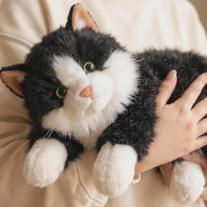 May include: A plush cat toy featuring black and white fur, green eyes, and a pink nose. The stuffed animal has white paws and a white chest, with black fur covering the rest of its body, including its ears which have light tan inner details.