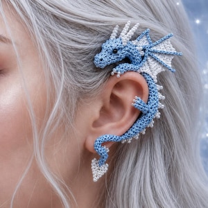 May include: A crochet ear cuff shaped like a dragon, featuring light blue yarn with white accents on the wings, crest, and tail tip. The design includes small white beads along the body and wings, with a dark bead for the dragon's eye.