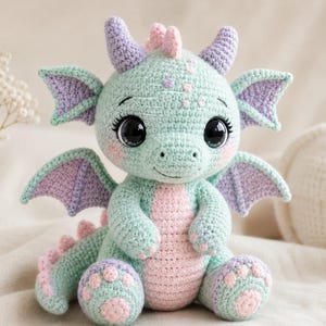 May include: A crocheted dragon plushie in pastel colors. The dragon features a mint green body, lavender horns and wings, and a pink belly and accents. It has large, expressive eyes and is sitting on a soft surface.