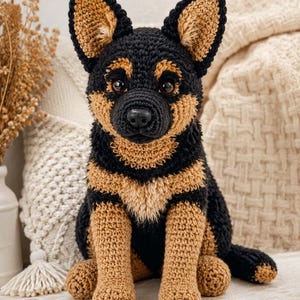 Könnte beinhalten: Ein handgehäkelter Hund in Schwarz und Hellbraun. Der Hund hat große, aufrechte Ohren, eine schwarze Nase und braune Augen. Die Häkelstiche sind sichtbar und verleihen dem Hund ein strukturiertes Aussehen. Der Hund sitzt aufrecht.