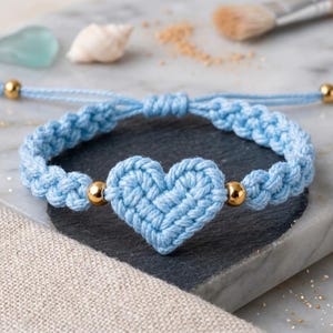 May include: A light blue macrame bracelet with a woven heart-shaped centerpiece and gold-colored beads. The bracelet is adjustable and sits on a marble surface. The bracelet is a handmade jewelry item.