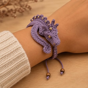 May include: A purple crochet bracelet shaped like a dragon, featuring small gold-colored beads along the spine and head. The bracelet has a flexible band design with two dangling strings at the end, each finished with a small brown wooden bead.