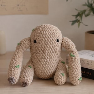 Puede incluir: Un peluche beige tejido a ganchillo con cuatro brazos y un cuerpo redondeado. El juguete tiene pequeños ojos negros y detalles verdes en los brazos. El peluche está sobre una superficie de madera clara.