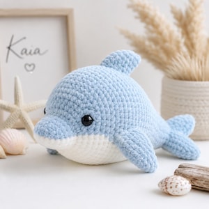 Puede incluir: Un peluche de delfín tejido en crochet azul claro con vientre blanco y ojos negros. El animal de peluche hecho a mano se exhibe con una impresión enmarcada que dice "Kaia" y un símbolo de corazón, junto con conchas marinas y elementos decorativos.