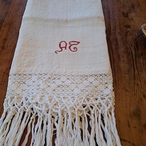 May include: An off-white rectangular linen textile featuring a red embroidered monogram of the letters "AS" in the center. The lower section displays a wide band of intricate white crocheted lace trim finished with long, knotted fringe tassels.