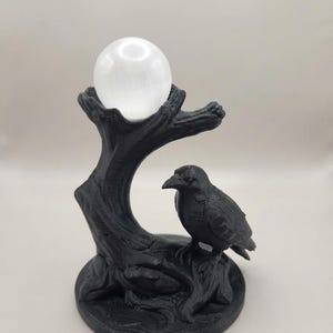 May include: A black 3D-printed sculpture featuring a raven perched on the base of a tree stump. The stump has a hollowed top section that holds a smooth, spherical white crystal ball. The entire object is set on a circular black base.
