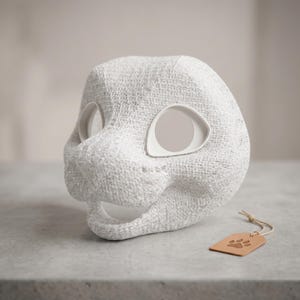 Puede incluir: Una máscara blanca con textura en forma de animal, con grandes aberturas para los ojos y la boca abierta. La máscara presenta un acabado superficial rugoso y poroso, acompañada de una pequeña etiqueta rectangular marrón claro con una huella.