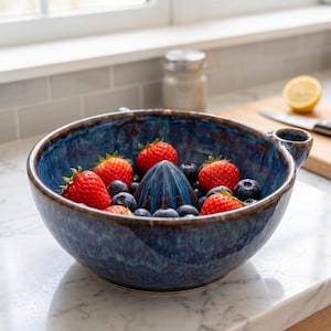 Puede incluir: Un exprimidor de cítricos de cerámica azul profundo con un cono central, que presenta un acabado de esmalte brillante y moteado. El cuenco está lleno de fresas rojas frescas y arándanos azul oscuro, mostrando un diseño de utensilio funcional.