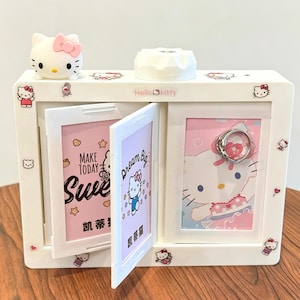Puede incluir: Una caja de joyería Hello Kitty blanca con una figura de Hello Kitty en la parte superior. La caja tiene dos puertas con bisagras con imágenes rosas y blancas en el interior. Una puerta tiene el texto "Make Today Sweet" y la otra dice "Dream Big". Un anillo se muestra en el interior.